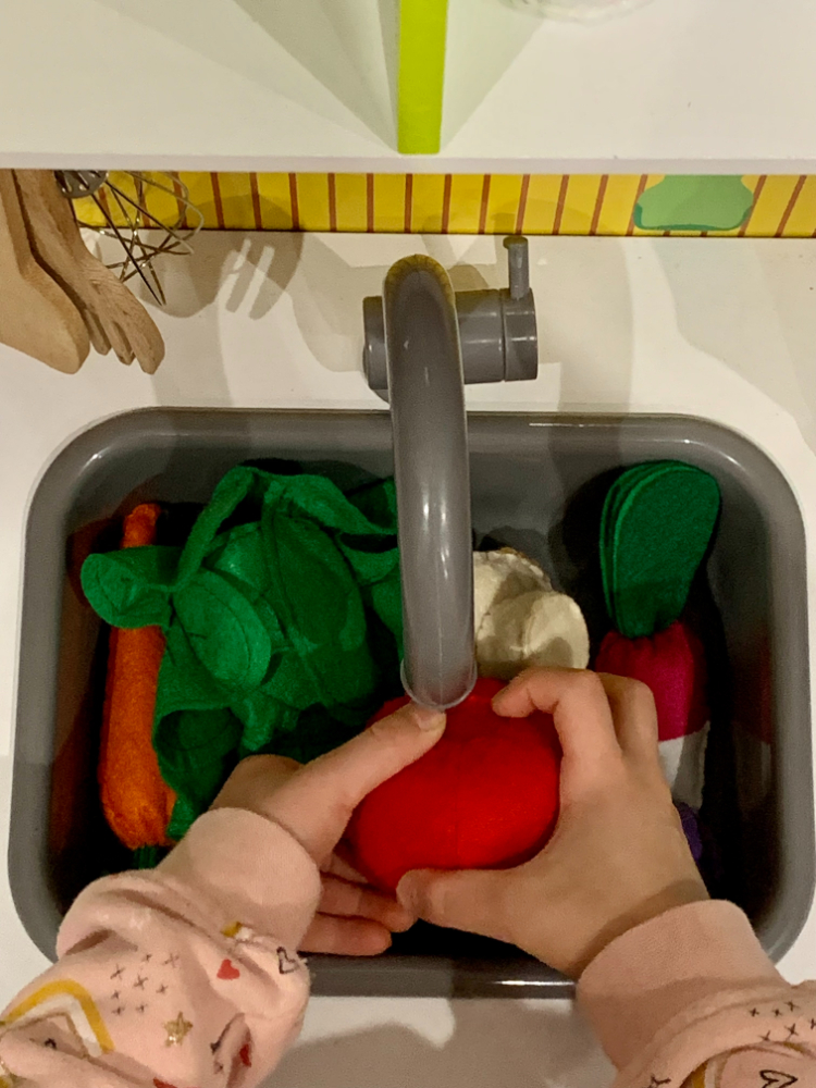 Enfant jouant à la dînette en feutrine, en imitant le geste de laver des légumes dans sa cuisine.