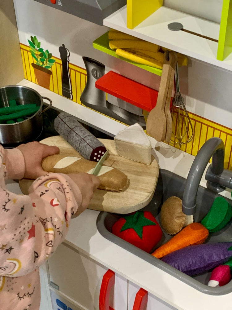 Enfant jouant à la dînette en feutrine, en imitant le geste de couper une baguette dans sa cuisine.